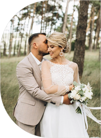 Pan młody w beżowym garniturze całuje pannę młodą w czoło w lesie. Panna młoda w koronkowej sukni trzyma bukiet i uśmiecha się delikatnie. Scena jest spokojna i romantyczna.