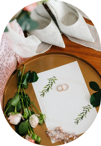 Płaskie ułożenie eleganckich białych koronkowych butów, dwóch złotych pierścionków, delikatnych zielonych liści, różowych róż i koronki na drewnianej powierzchni, przywodzące na myśl romantyczną atmosferę